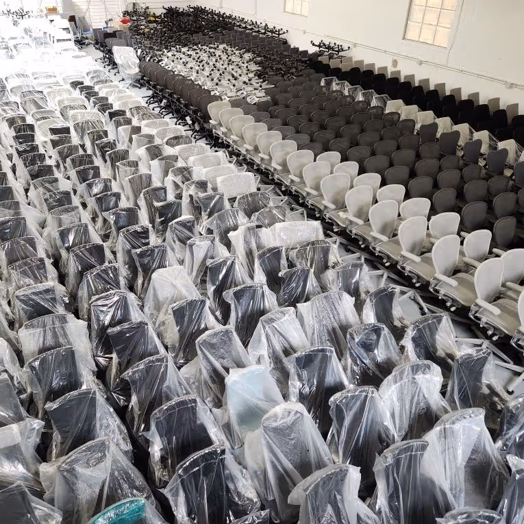 Large warehouse filled with rows of office chairs, some covered in plastic wrap, in black and white colors.
